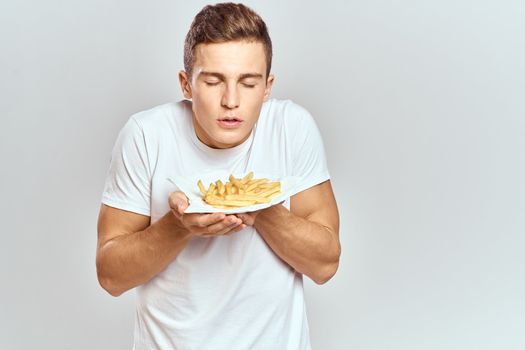 young guy with french fries in a box and in a white t-shirt on a light background cropped view. High quality photo