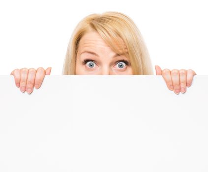 Young woman with surprised eyes peeking out from behind billboard paper poster. Businesswoman holding big white banner, isolated on white background.