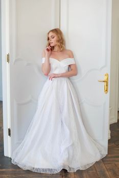 A bride in a chic wedding dress stands near the door and looks to the side thoughtfully