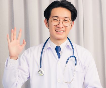 Portrait of Happy Asian young doctor handsome man smiling in uniform with stethoscope talking online video conference call or facetime raise his hand to say hello, healthcare medicine concept