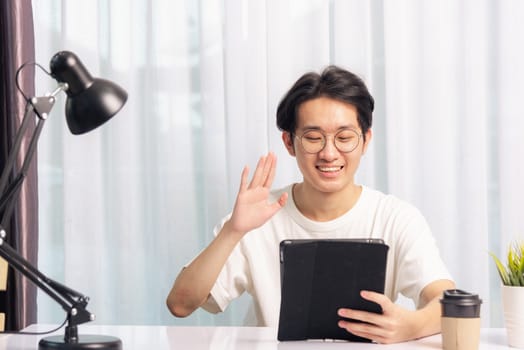 Happy Asian young business handsome man work from home office wear glasses, t-shirt comfortable he smiling and using a black modern smart digital tablet computer raise hand to say hello team on desk