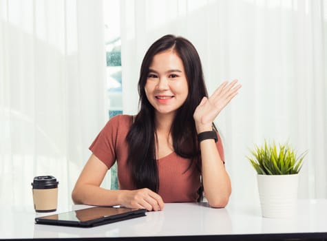 Work from home, Asian business young beautiful woman smiling sitting on desk workspace raise hand say hello communicating with team video chat or conferencing on digital tablet computer at home office