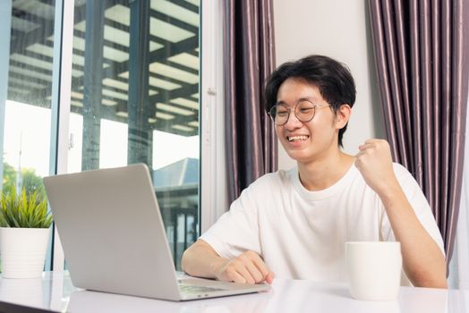 Happy Asian young business handsome man smile work from home office wear glasses, t-shirt comfortable exciting good news successful winner raises hand yes gesture with laptop computer on desk at home