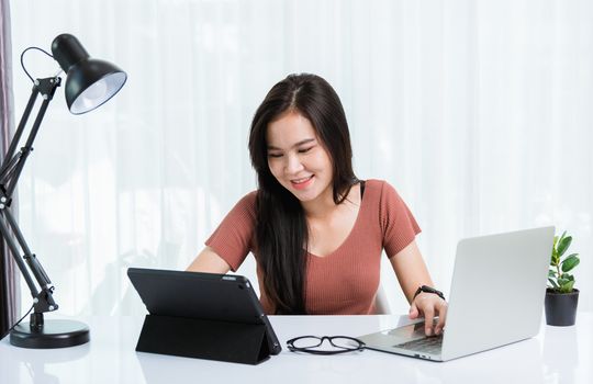 Work from home, Smiling Happy Asian business young beautiful woman sitting on desk workspace use modern smart tablet and laptop computer technology video call conferencing to team at home office