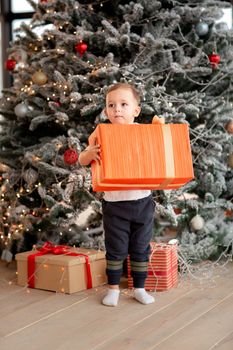 Cheerful cute baby boy with gifts. Kid holding big present and having fun near tree in the morning. Merry Christmas and Happy Holidays concept