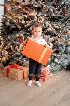 Cheerful cute baby boy with gifts. Kid holding big present and having fun near tree in the morning. Merry Christmas and Happy Holidays concept