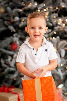 Cheerful cute baby boy with gifts. Kid holding big present and having fun near tree in the morning. Merry Christmas and Happy Holidays concept
