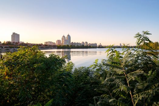 High Obolon buildings near the Dnieper river in Kiev, Ukraine. Blue clear sky and reflection in the water. Various plant and vegetation in the foreground