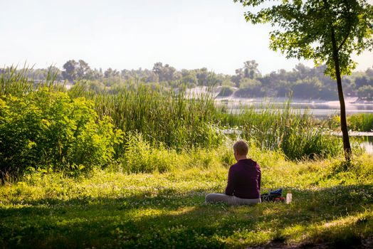 Kiev / Ukraine - July 16, 2020 -  A woman is meditating, sitting in the grass in front of the river in the morning, just after sunrise.