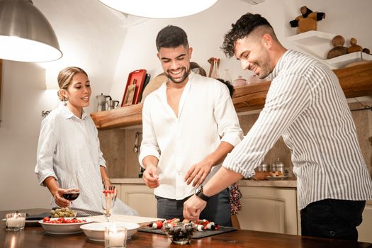 Group of three friends at home preparing savory snacks for the aperitif - Two young man and a young woman smiling in the kitchen waiting for the happy hour