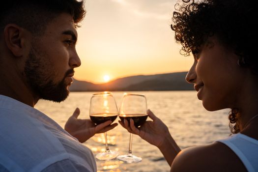 Romantic close-up of a mixed race couple toast with glasses of red wine crossed by the light of the setting sun on a lake