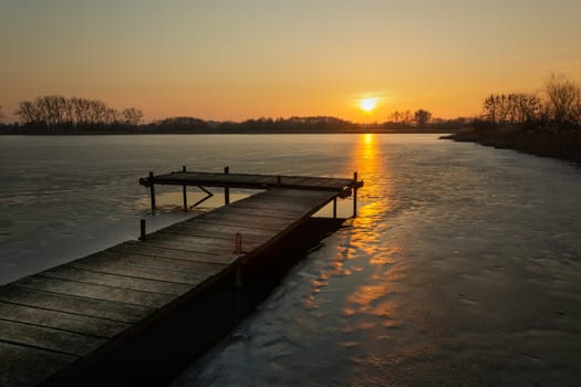 Beautiful evening view of the wooden platform from the planks on the frozen lake at orange sunset.