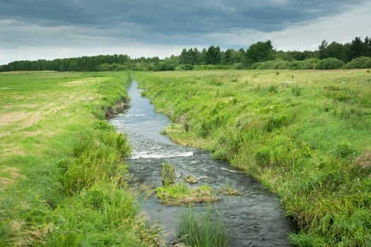 A small river flowing through meadows and clouds in the sky, summer view