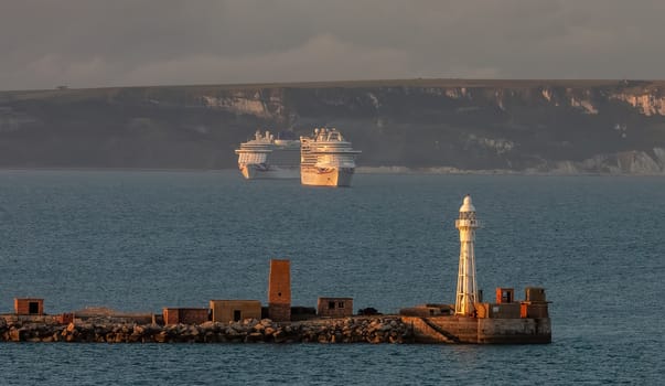 Portland, United Kingdom - July 1, 2020: Portland harbour lighthouse with two P&O cruiseships anchored in the distance in Weymouth Bay at sunset.