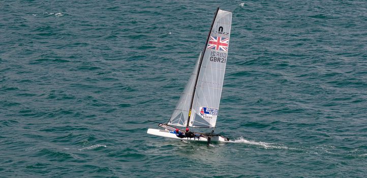 Portland harbour, United Kingdom - July 2, 2020: High Angle aerial panoramic shot of racing catamaran of the British Sailing Team. Two sailors on it wearing red helmets, British flag on the sails.