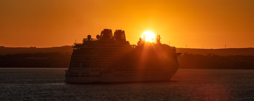 Weymouth Bay, United Kingdom - July 10, 2020: Amazing panoramic shot of P&O cruise ship Britannia anchored in Weymouth Bay at sunset. Sun setting down right above the ship casting orange color on it