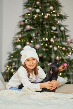 Happy childhood, Christmas magic fairy tale. A little girl is laughing with her friend, a dachshund dog, near the Christmas tree.