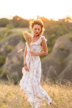 Beautiful tender girl in a white sundress walks at sunset in a field with a spikelet bouquet