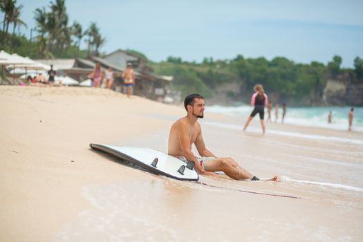 The guy is resting on a sandy tropical beach, after riding a surf. Healthy active lifestyle in summer vocation.