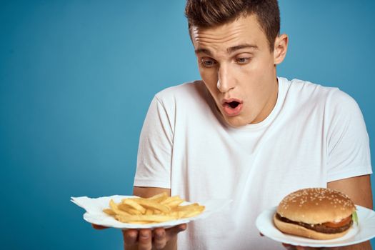 young guy with fries and hamburger on blue background interested look emotions fast food calories cropped view Copy Space. High quality photo