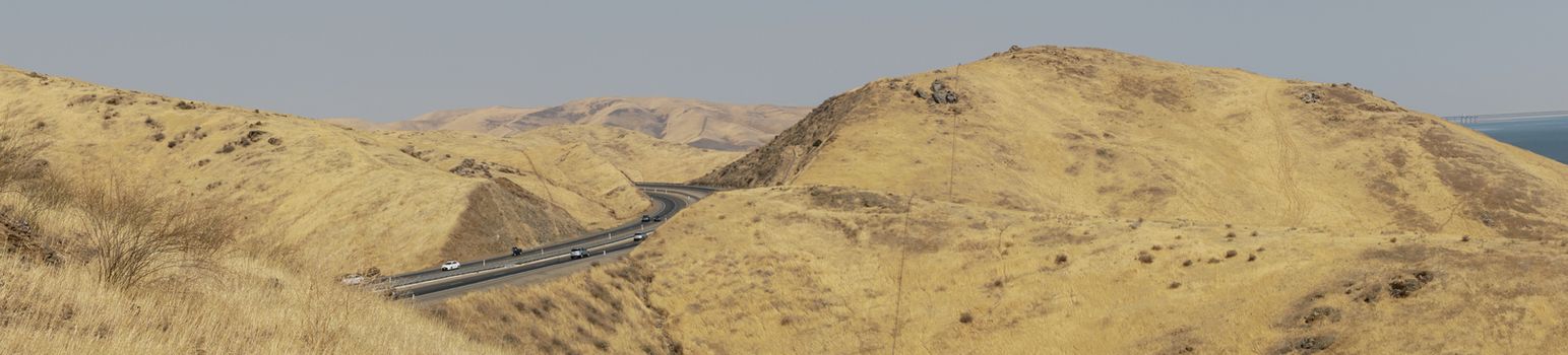 Panoramic view of the freeway road with cars crossing the the San Luis Reservoir valleys during dry season, San Luis Creek in the eastern slopes of the Diablo Range of Merced County, California. USA
