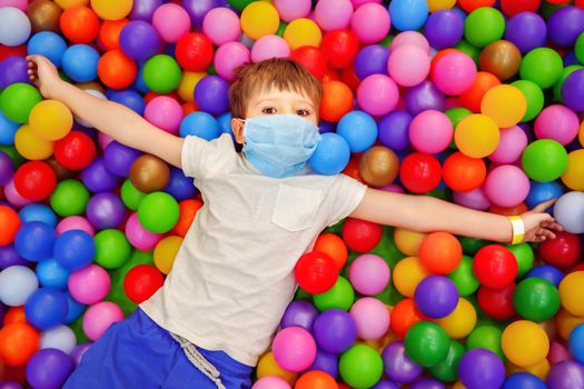 Cute caucasian boy lying in dry pool with plastic balls. Boy wearing protective medical mask at entertainment center. Leisure activities indoors during quarantine. Illness prevention concept. Top view