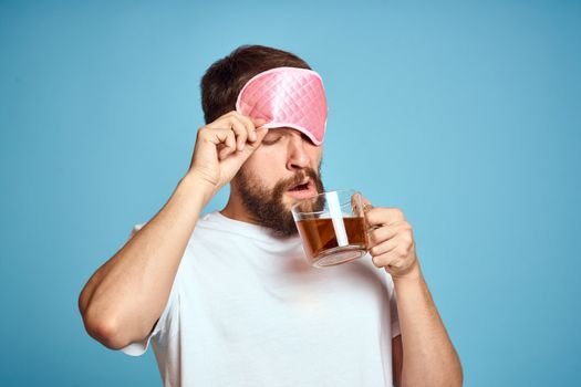 Man with pink sleep mask on his face and a cup of tea in his hands cropped view close-up blue background. High quality photo