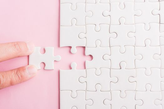 Top view flat lay of hand-holding last piece white paper jigsaw puzzle game last pieces put to place for solve problem complete mission, studio shot on a pink background, quiz calculation concept