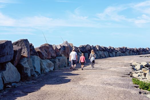 A family group of two young people and two older persons, unidentified, walking slowing along a marina breakwater wall on a bright sunny day with blue sky and water.