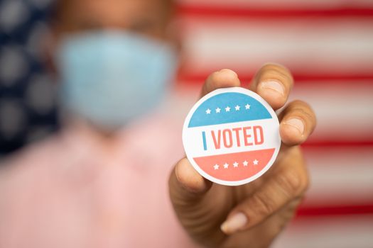 Close up shot man in medical mask showing I voted Sticker and putting on shirt with US flag as background - concept of US election