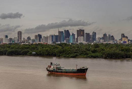 Bangkok, thailand - Sep 20, 2020 : A cargo ship parked in the middle of the river and in front of a green forest with skyscrapers at evening by the Chao Phraya River.