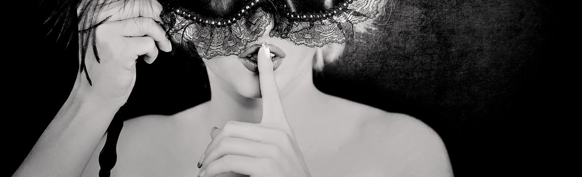 Close up in black and white of the lower face of a beautiful young woman wearing a lacey mask and with her finger to her lips