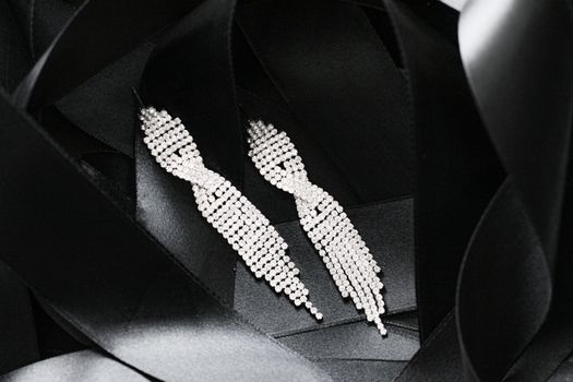 Luxury diamond earrings on black silk ribbon as background, jewelry and fashion brands