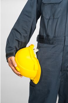 Worker standing in blue coverall holding yellow hardhat isolated on gray background