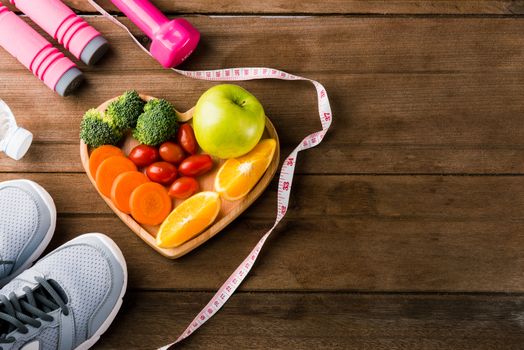 Top view of fresh fruits and vegetables in heart plate wood (apple, carrot, tomato, orange, broccoli) and dumbbells, sport shoes sports equipment on wooden table, Healthy lifestyle diet food concept