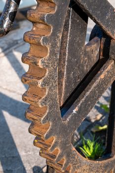 A large iron cog of a gear with rust forming on its outer prongs is seen up close outdoors, showing texture and detail.