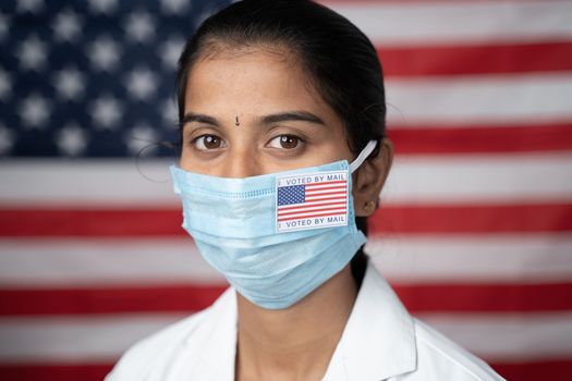 Close up face of Girl with I voted by main sticker on her medical mask with US flag as background - Concept of mail in voting in US election