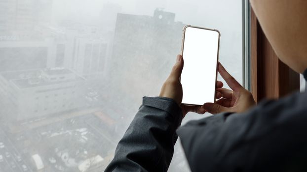 Woman hand holding mobile phone with blank white screen mockup near window at outside a snowy.
