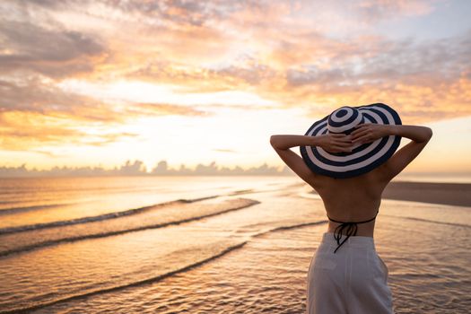 Woman in top bikini and white long pant wearing hat on the beach with a beautiful sunrise or sunset in background.
