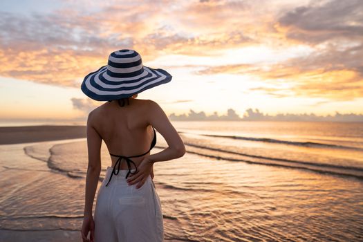 Woman in top bikini and white long pant wearing hat on the beach with a beautiful sunrise or sunset in background.