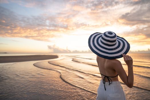 Woman in top bikini and white long pant wearing hat on the beach with a beautiful sunrise or sunset in background.