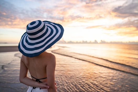 Woman in top bikini and white long pant wearing hat on the beach with a beautiful sunrise or sunset in background.