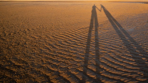 The shadow of a very long couple holding hand on a sand beach surface.