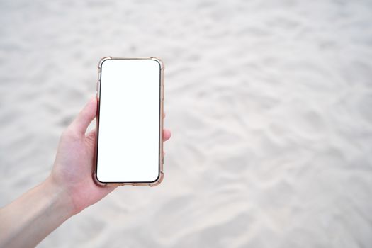 Woman hand holding mobile phone with blank white screen mockup on the beach.
