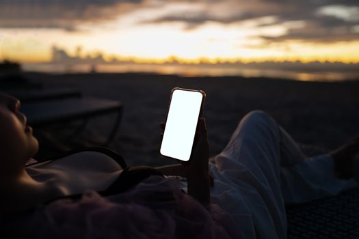 Woman hand holding mobile phone with blank white screen mockup on the beach in the morning.