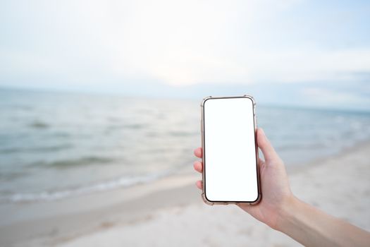 Woman hand holding mobile phone with blank white screen mockup on the beach.