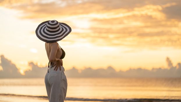 Woman in top bikini and white long pant wearing hat on the beach with a beautiful sunrise or sunset in background.