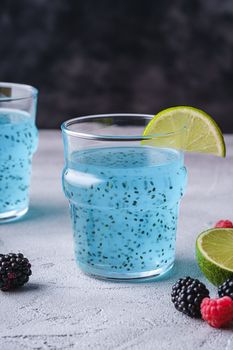 Tasty blue colored cocktail drink with basil chia seeds, citrus lime slice, raspberry and blackberry berries in two glass, healthy summer beverage, stone concrete background, angle view