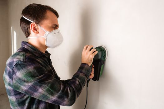 Man in protective mask working with electric sander to smooth plaster wall surface, room renovation concept