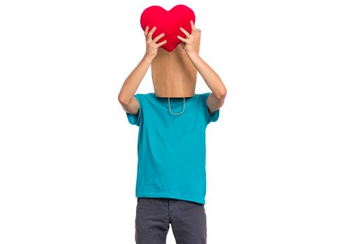 Valentines Day concept. Teen boy with paper bag over his head holds red heart, isolated on white background. Boy holding symbol of love, family, hope. Teenager cover head with bag posing in studio.